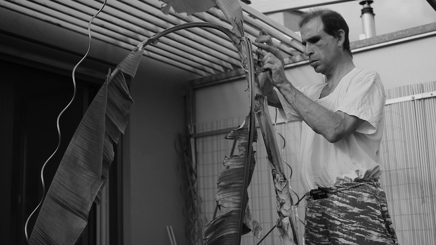 A part of the film “Imprint”. A man is touching the banana tree on his balcony. His eyes are closed.