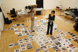 Photo of two women standing in an office room with picture pages spread on the floor around them, the person on the right is also holding picture pages in her hand, both are looking at the camera