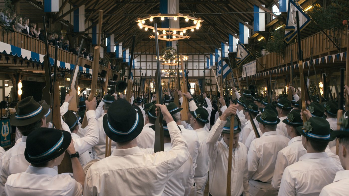 photo of a gathering of hunters in white shirts standing together in a large wooden hall with green hunter hats and holding their guns in the air, blue and white flags and garlands hanging everywhere