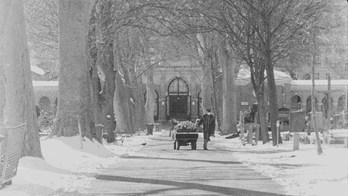 Erde (16mm-practice) by Lukas Ellmer and Gabriel Monthaler. AUT 2022, B/W, 5 min black and white shot of a cemetery employee driving a coffin decorated with flowers along an avenue at the cemetery covered with snow
