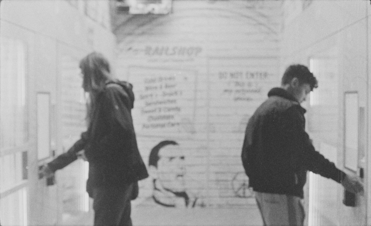 Railshop (16mm-practice) by Konrad Behr and Johannes Lehnen. AUT 2022, color, 7 min black and white photo of two people standing with their backs to each other in front of vending machines, in the background is the "Railshop" train