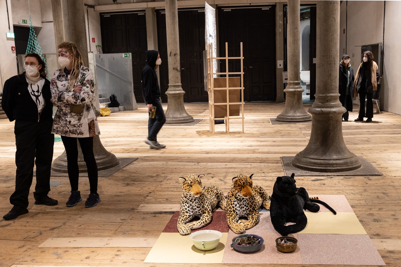 Several people wearing FFP2 masks stand in a room with columns and wooden floor. In the foreground you can see an artistic work: 3 stuffed wild cats, 2 leopards a panther lying on the floor in front of feeding bowls in the same position.