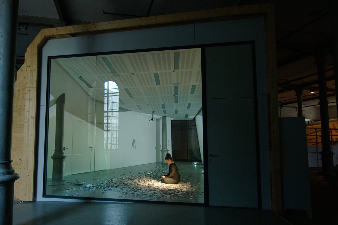 A performer sits cross-legged in a room behind a glass wall and tears up photos. The snippets are spread around her, she wears black clothes, her hair is pinned up. The surrounding space is reflected in the glass.