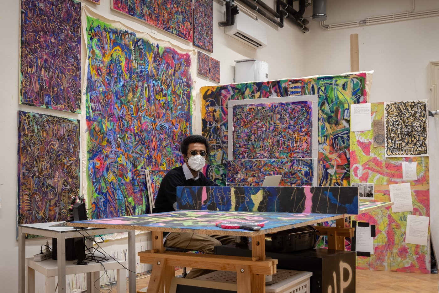 The artist sits behind his work table with mask and sunglasses. He is surrounded by his works, a multitude of small-scale, colorful and abstract paintings and drawings.