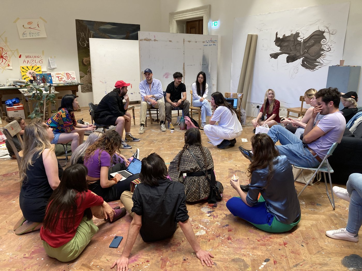Class meeting of the department of figurative painting.18 Students of the class are gathered in room E 18 at Schillerplatz.They are discussing and exchanging ideas.  In the background are works in progress of Gregor Hagenauer and Bergur Nordal.