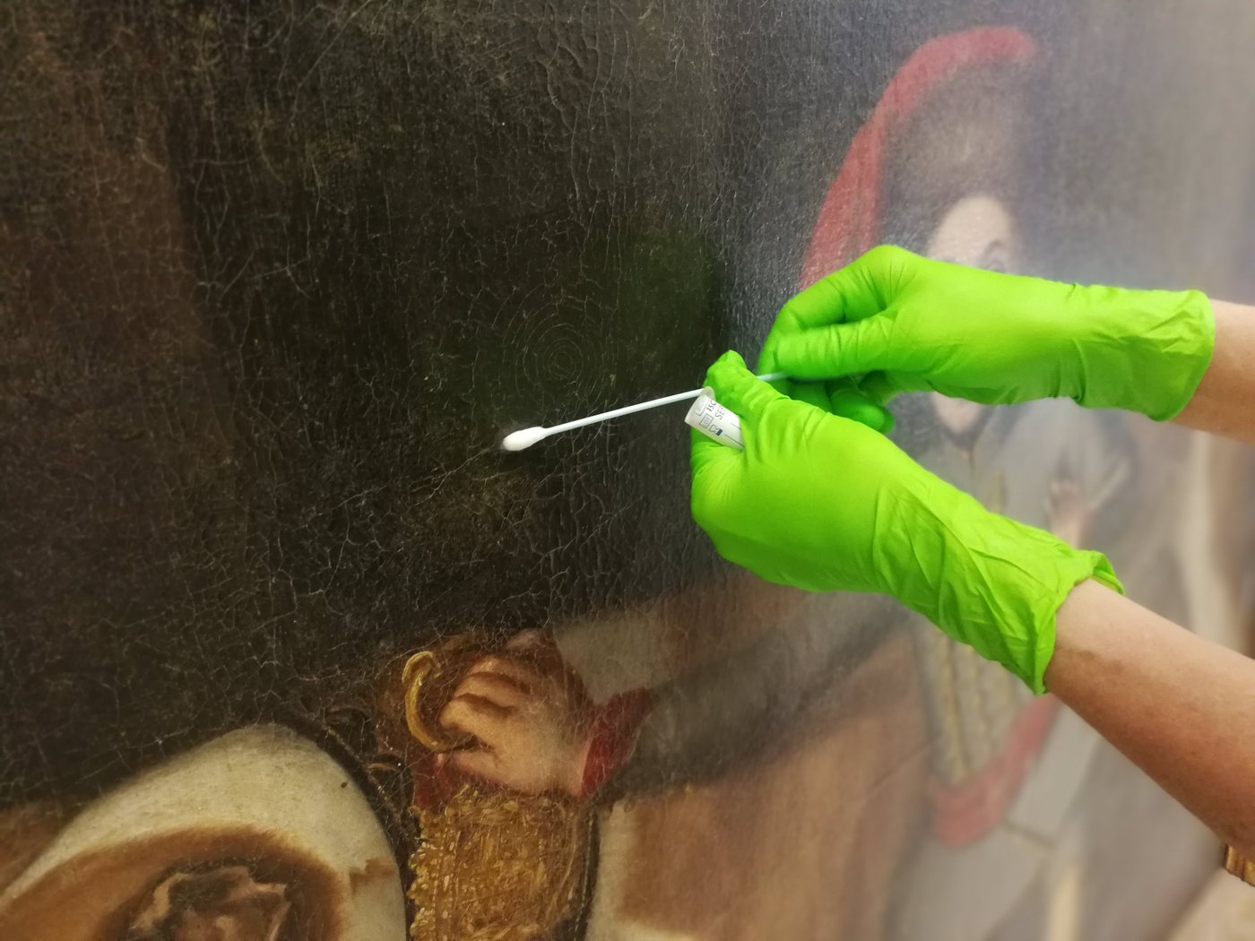 Gloved hands hold a sample tube between thumb and forefinger while simultaneously stroking the surface of an oil painting with a cotton swab.