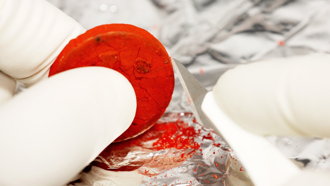 Close-up of gloved hands scraping tiny pieces from a bright red tablet of watercolour paint with a scalpel. The fragments are collected on aluminium foil.
