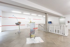 view of a room with exhibition items in front, some city maps on the floor and on the wall in the back, also two lines of red stripes on the wall