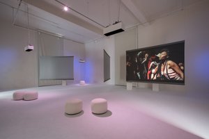 white room with white round stools, in the background several screens hanging on wires, on one screen is projected a movie with a black woman in the picture