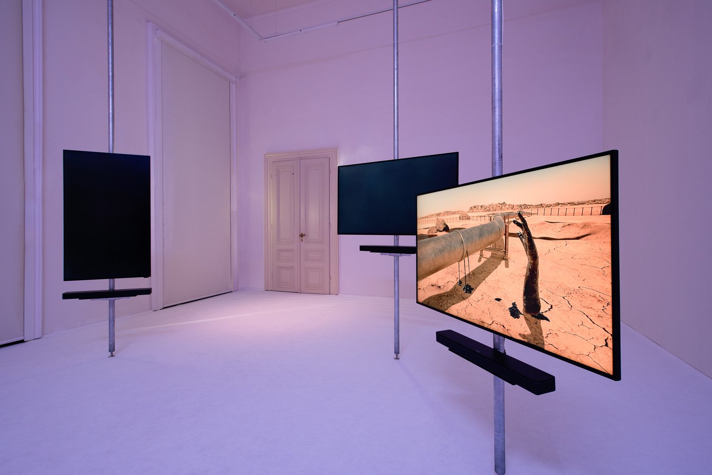 white room with three screens mounted on poles and distributed in the room, two screens are black, on the third one there is a picture of a desert landscape with steel pipes and a black oily hand sticking out of the floor, in the background there is a white door