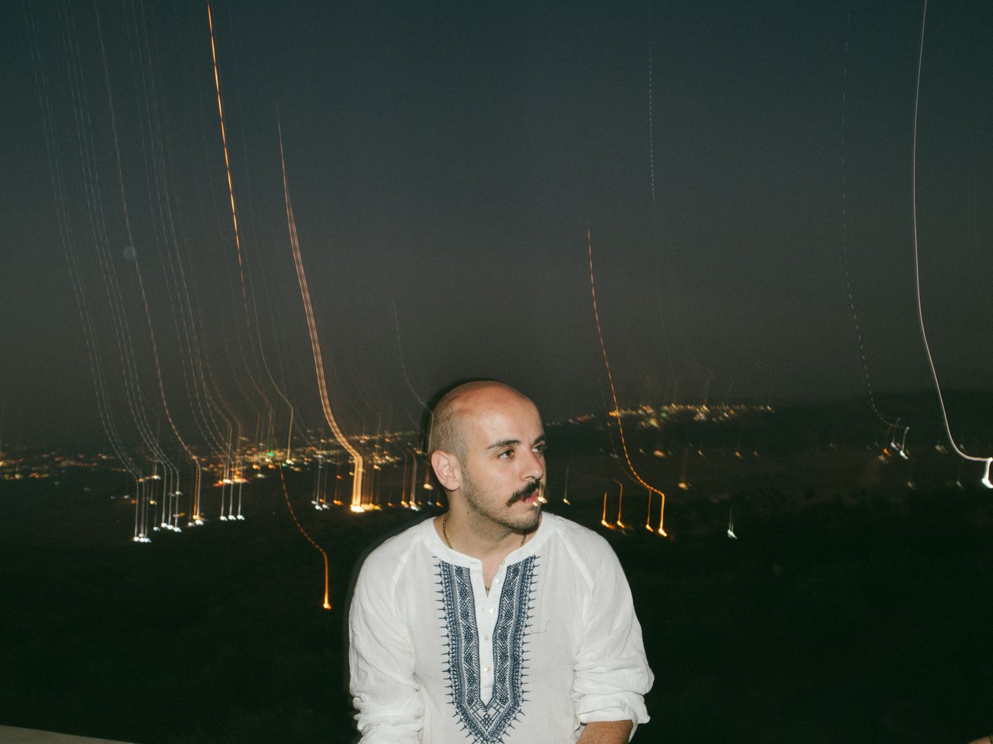 Half portrait of the artist, person with shaved hair, black moustache and three days beard on cheeks and chin in white, collarless shirt; in the background blurred lights of a city view at night;