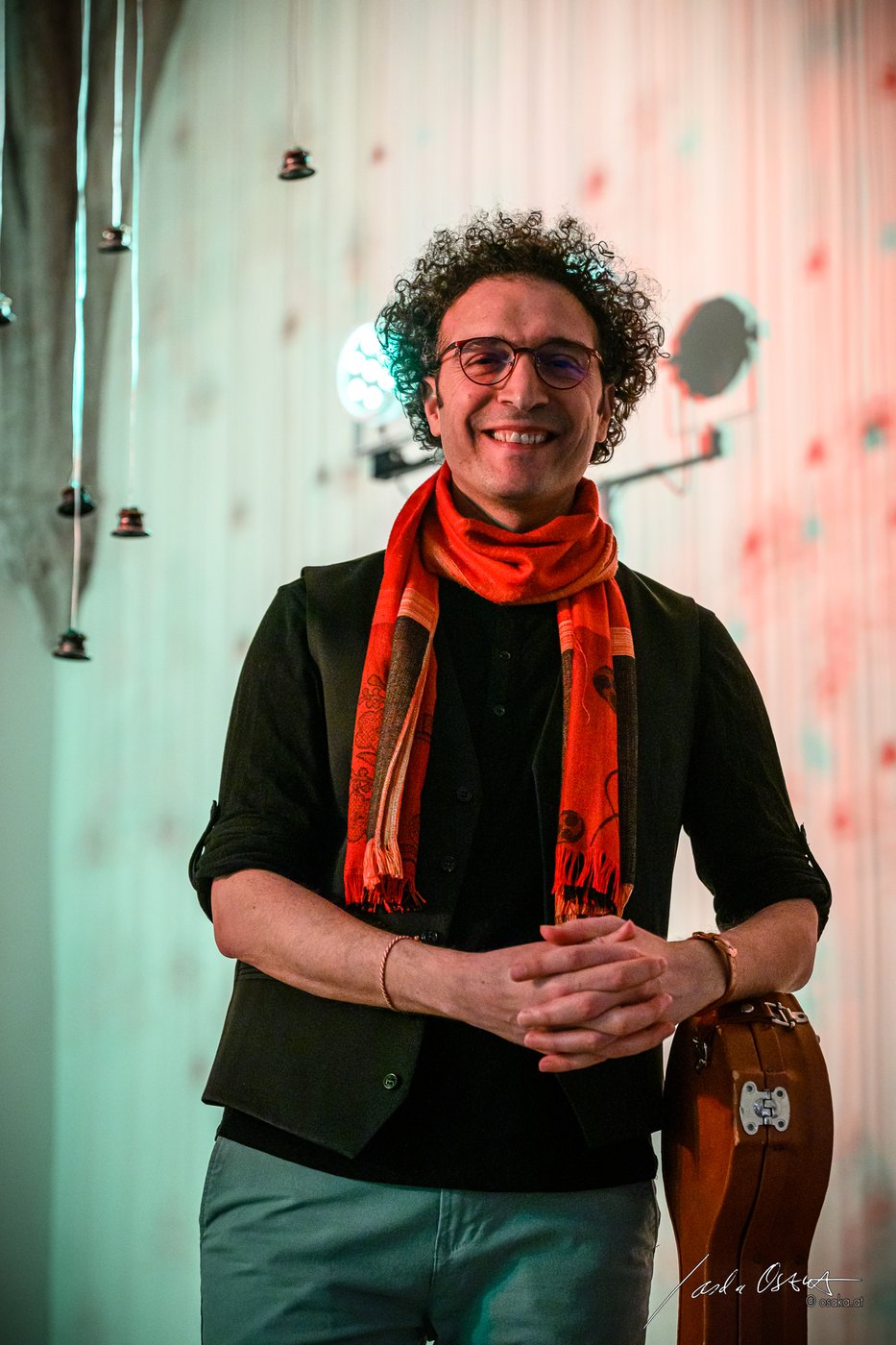 Musician standing, half portrait, leaning against a guitar case; green trousers, black shirt with black jelly over it and red scarf around his neck; in the background small bells hanging from the ceiling on ribbons;