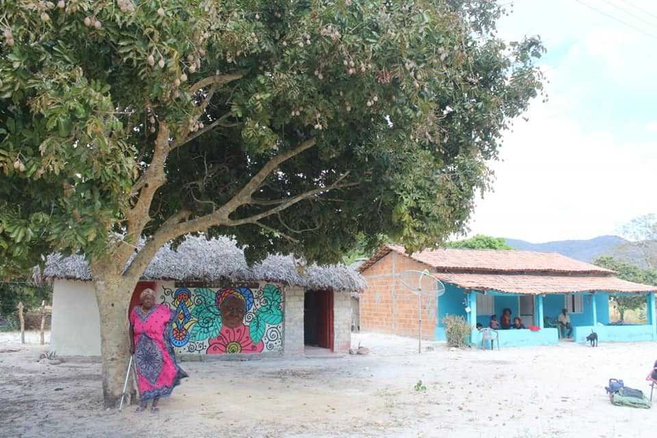 African woman in colorful dress in front of two houses which are also colorfully painted.