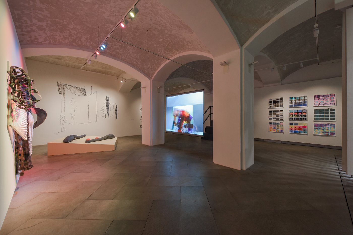 In the basement with vaulted ceiling you can see various works. An expansive wall object, a wall drawing, textile objects on a slanted pedestal, a film screen and 6 framed serial paintings.