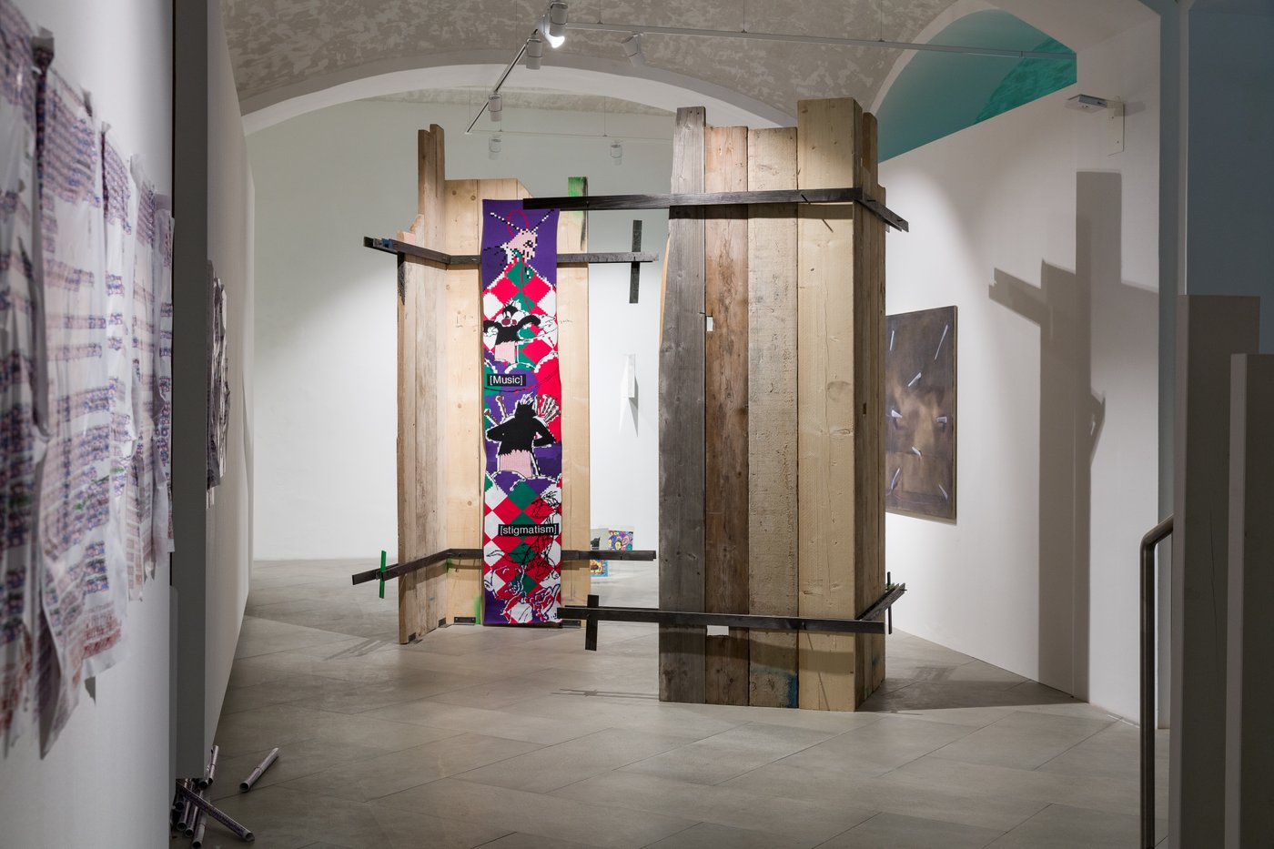 In the room there are two wooden sculptures made of planks, each forming a kind of corner of the room and facing each other. Inside one of the corners hangs a purple, red, green, white banner with the inscription "music", "Stigmatism".
