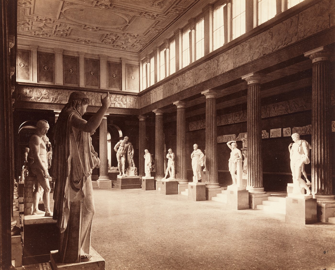 A historical photograph in sepia colors showing a large room with a row of columns between which are Greek sculptures on pedestals, above the columns a picture frieze and high upper windows through which light shines in