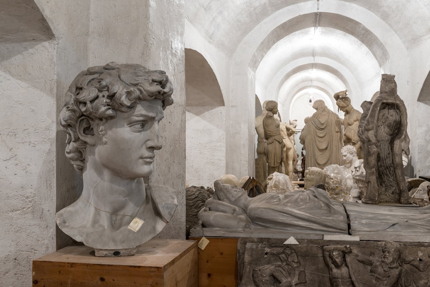 Photo of a room with vaults in which several Greek sculptures in different poses are standing close to each other. In front is the head of a young man.