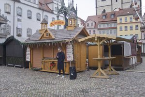 a man in a blue coat stands outdoors in front of a closed market with a suitcase