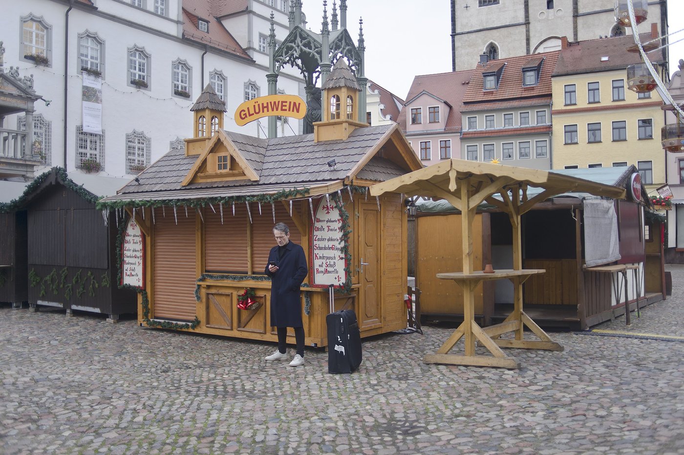 a man in a blue coat stands outdoors in front of a closed market with a suitcase
