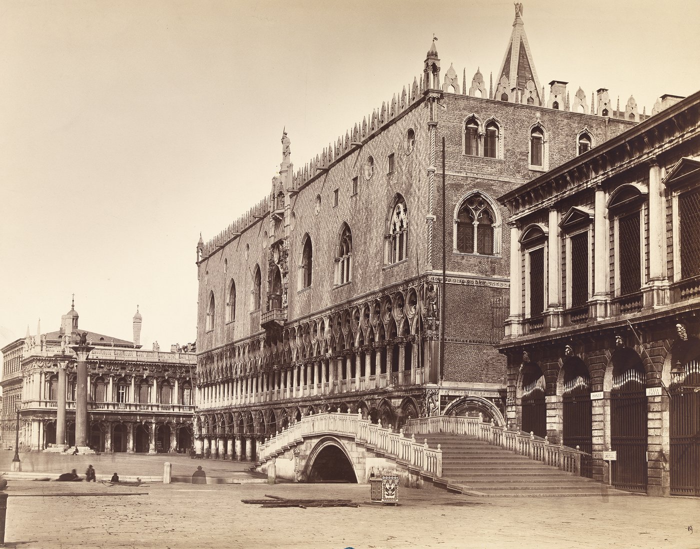 A black and white sepia photograph depicting the Doge's Palace and a small bridge. The architecture corresponds to the Venetian Gothic style and therefore shows divided windows and ornaments tapering towards the top, as well as a loggia-like passageway supported by columns and pillars. On the left side of the picture, a few people are slightly blurred.