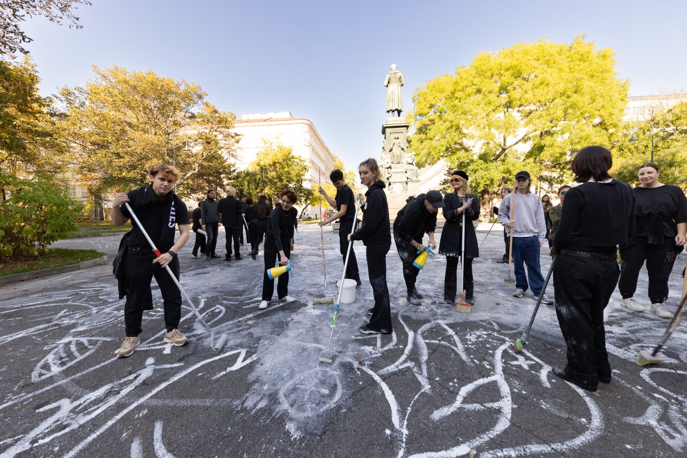 Several students working together to draw a chalk drawing with white paint and brooms on Schillerplatz.