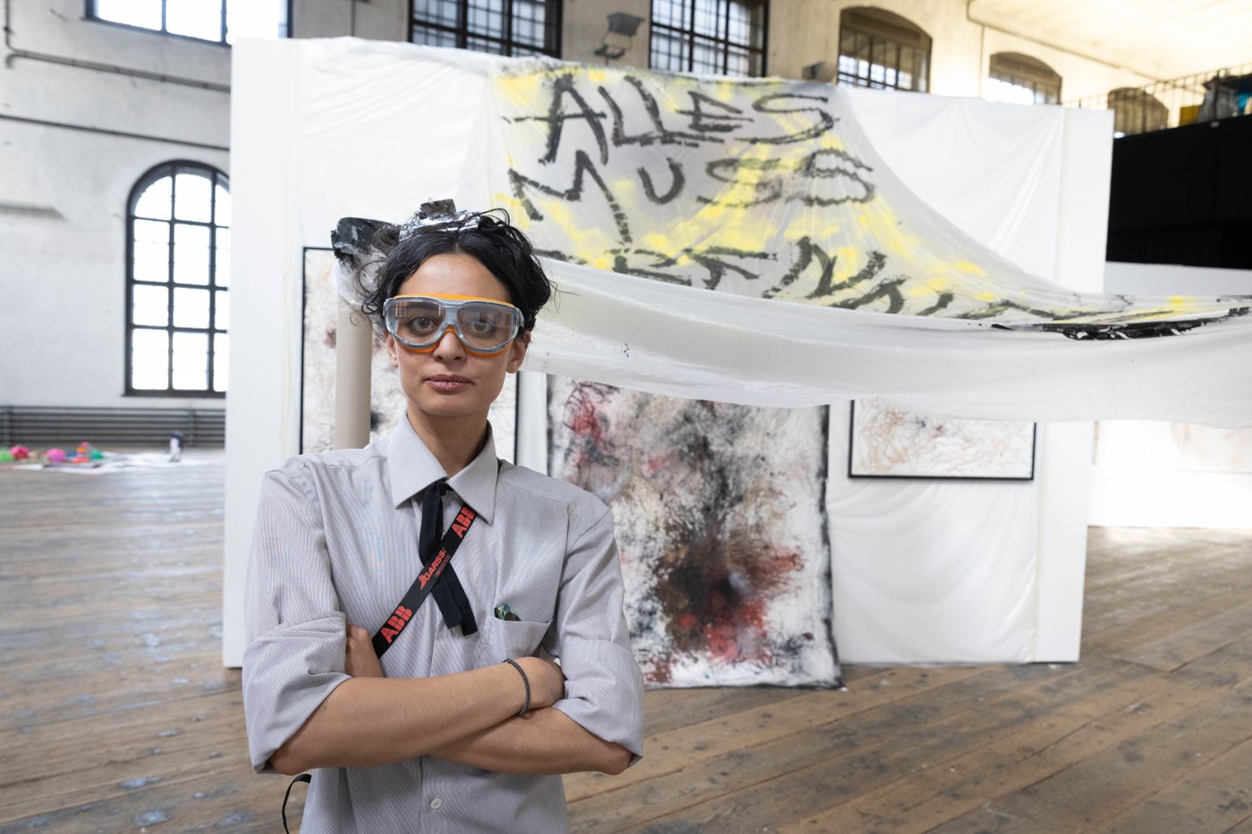 The artist in front of her work with secure glasses on and in the background there is a banner with the words "alles muss raus"