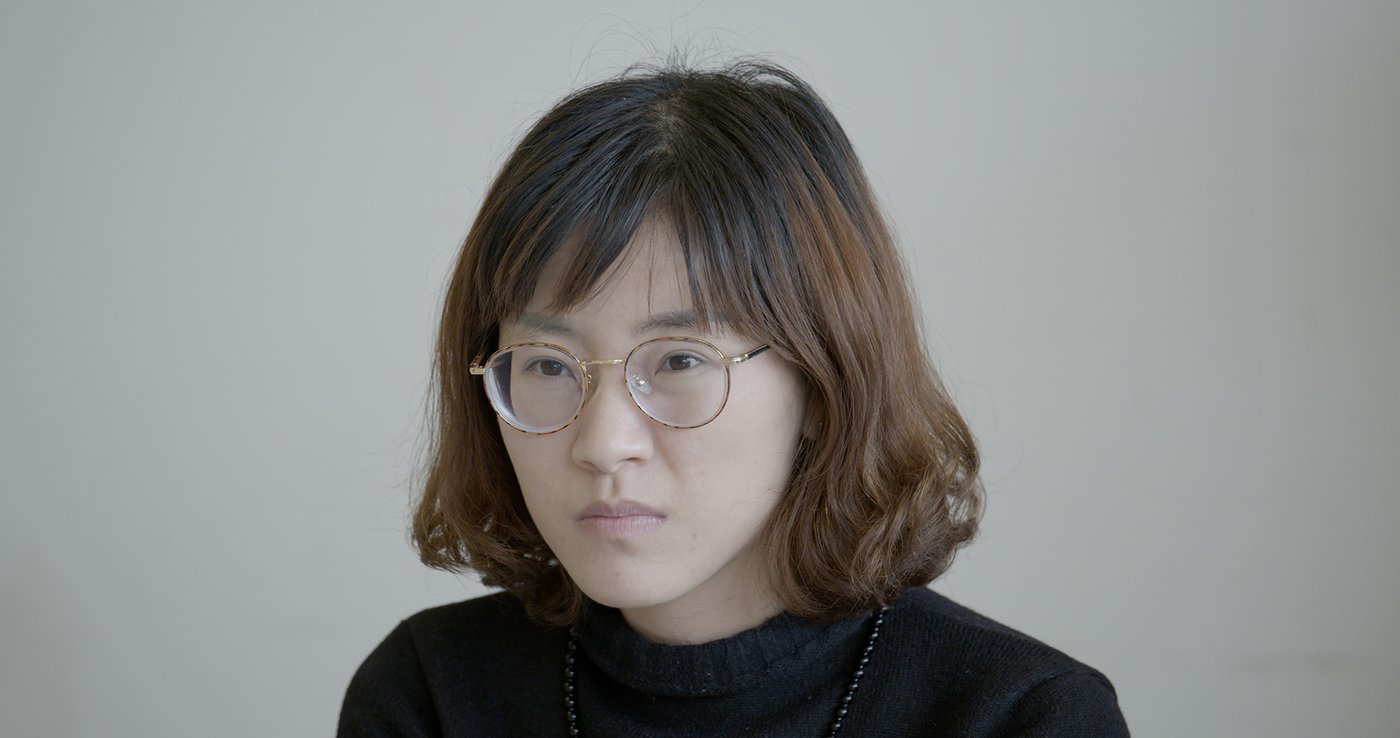 Photo of asian woman with shoulder length dark brown hair and round glasses, she stares past viewer with dogged look.