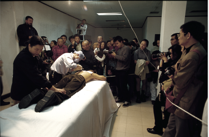 Photo of the performance Planting Grasses in the Fuck Off exhibition. A person is lying on their stomach on a platform. Another person with a mask and a device stands bent over the back of the lying person. Spectators stand next to them, some with cameras.
