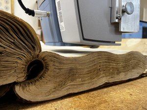 The image depicts the transportable Alpha II Fourier Transform Infrared Spectrometer manufactured by Bruker® instruments. The Alpha is positioned 1.5 cm above the pages of the manuscript. The manuscript shows a severe curl in the spine, making the spine a nearly perfect circle.