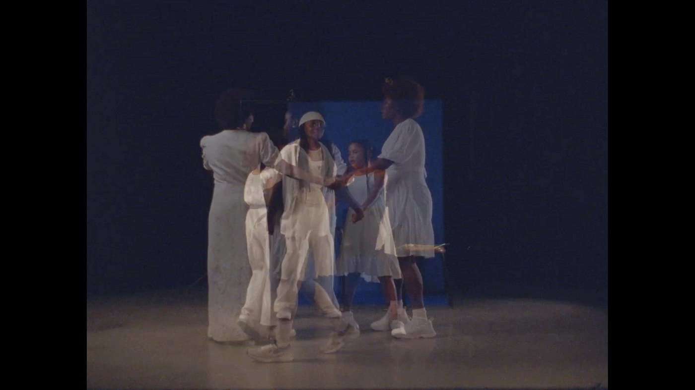 Five presons dressed in white walk in a circle while holding hands. Their bodies are transparent and a blue rectantle is visible in the background.