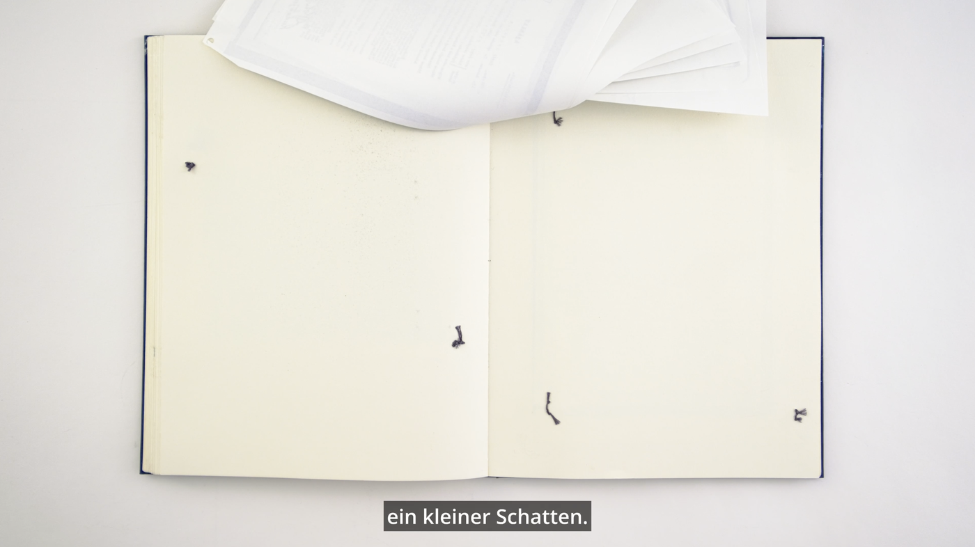 An open book with blank pages on a white background. Printed sheets lie upside down at the top of the book. Underneath the book is the inscription “ein kleiner Schatten” (a little shadow).