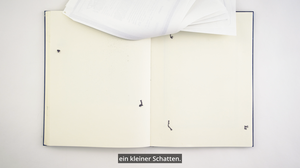 An open book with blank pages on a white background. Printed sheets lie upside down at the top of the book. Underneath the book is the inscription “ein kleiner Schatten” (a little shadow).