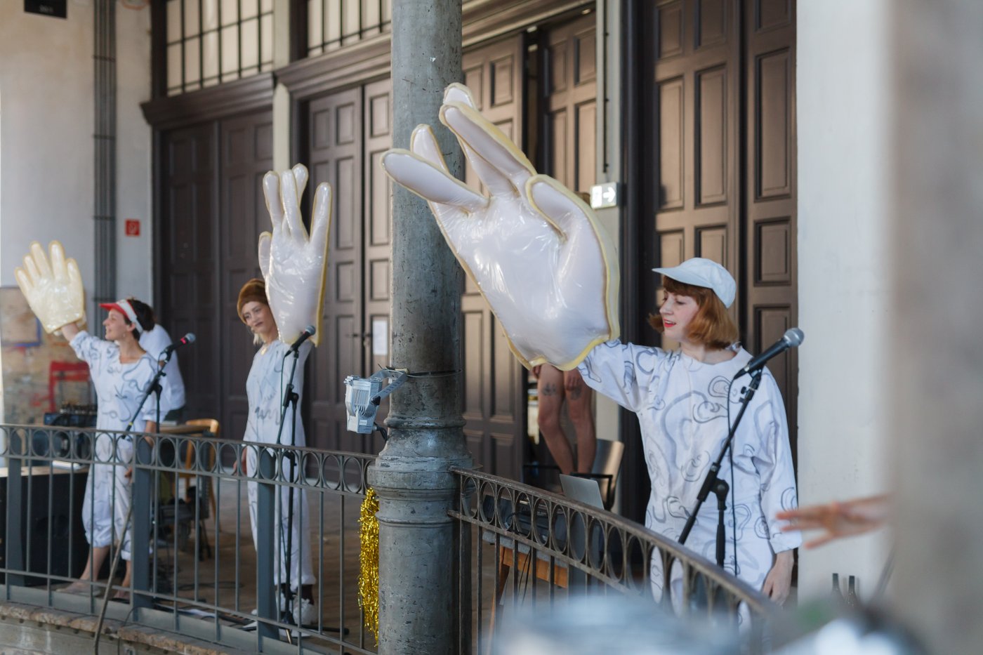 3 Women with oversized dummy hands wave to the audience as part of their performance in the Prospect Courtyard (large high space spanning 4 floors with galleries on each floor) during the 2019 graduation ceremony.