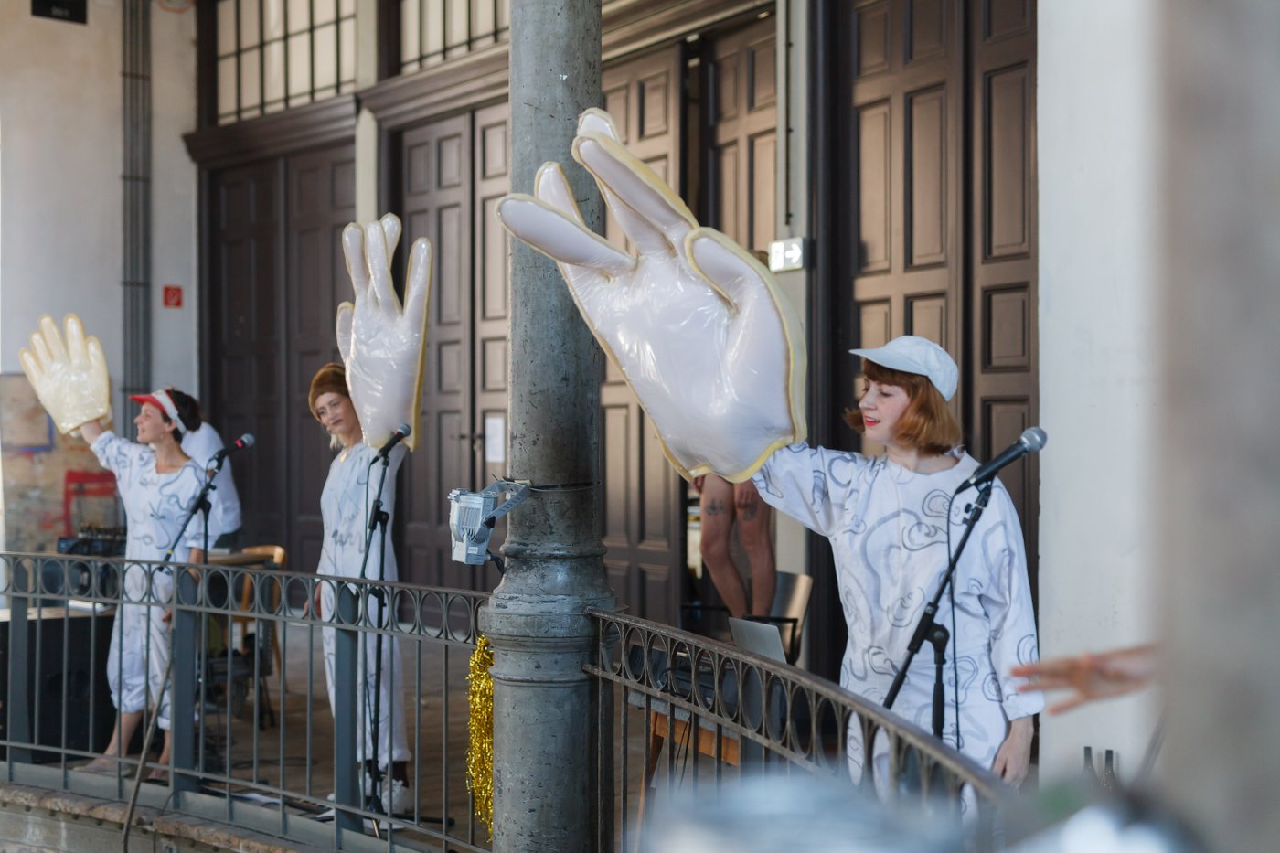 3 Women with oversized dummy hands wave to the audience as part of their performance in the Prospect Courtyard (large high space spanning 4 floors with galleries on each floor) during the 2019 graduation ceremony.