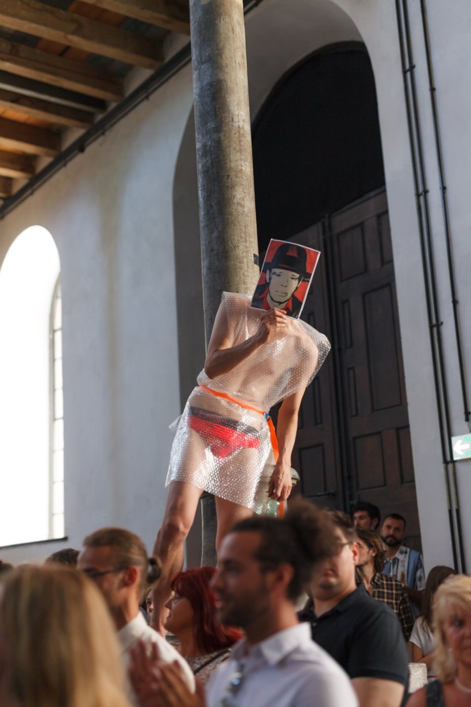 Artistic intervention at the graduation ceremony 2019. A person has wrapped his naked body with bubble wrap. You can see the red underpants, in front of the face he holds a portrait of Josef Beuys.