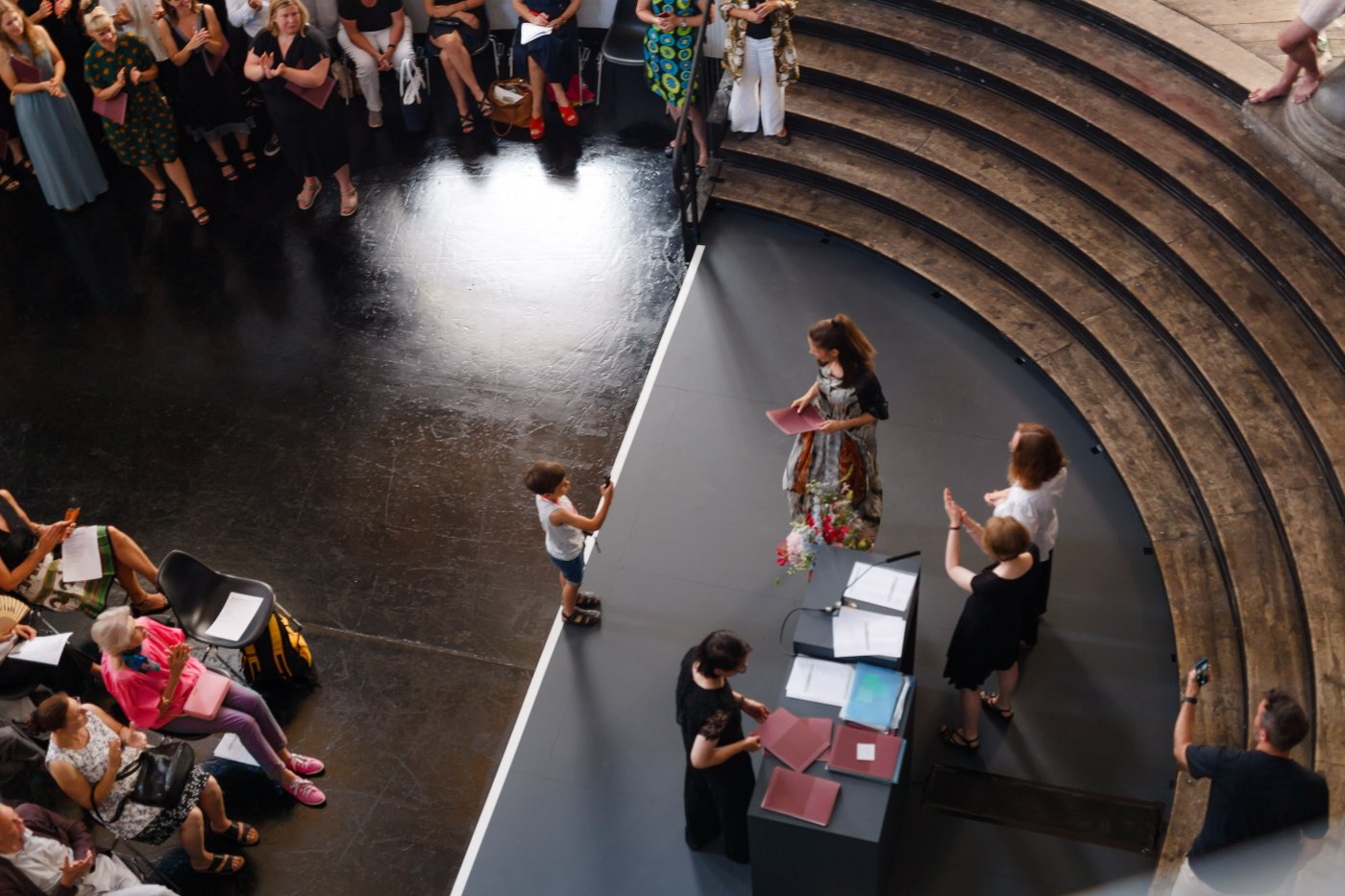 Presentation of certificates during the graduation ceremony 2019 in the Prospekthof: On stage, a graduand has a red folder in her hand, a little boy takes her picture, three other people applaud. The scene is seen from above, the people are standing on a kind of round stage surrounded by wooden stairs.