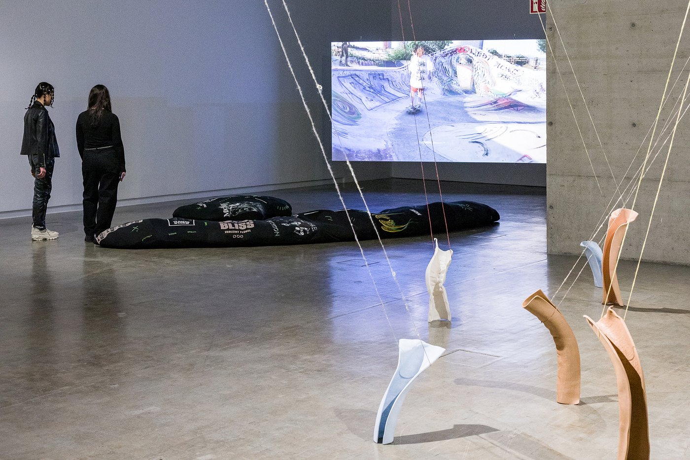 Exhibition view: on the gray floor in the foreground lies an exhibition object on the floor that looks like a large black beanbag - printed with terms - in front of it some colorful objects attached to strings, on the left of the picture two people in black clothes and in the background a screen with a video still on which a skateboard with a person can be seen.