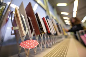 The somewhat blurry color photograph shows a long metal rack containing books of different shapes and colors. A small red cocktail umbrella with white dots is attached to the front left, and a woman looking at the books can be seen in the background.