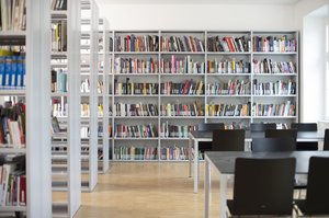 The color photograph shows a light-flooded room with a wooden floor and white walls. On the left and on the front wall are metal shelves filled with books, on the right are the working spaces with tables and chairs made of metal and black wood.