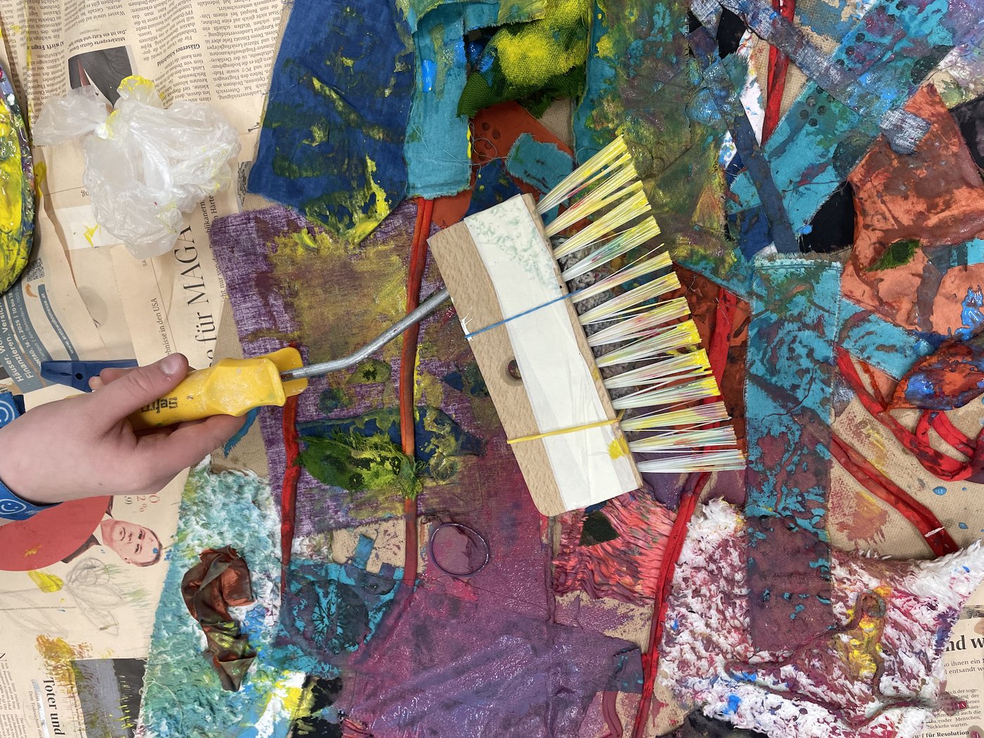 In the picture, you can see many bright colors painted on different fabrics and strings that are glued on top of each other. In the center, a hand is holding a brush with a yellow handle. In the background, there are areas of paint, pieces of paper, and parts of newspapers.