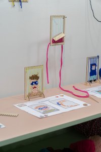Walldisplay showing numerous small weaving frames; one showing a handwoven completed portrait, the others are unfinished. A drawing with the design for the weavings is placed infront.