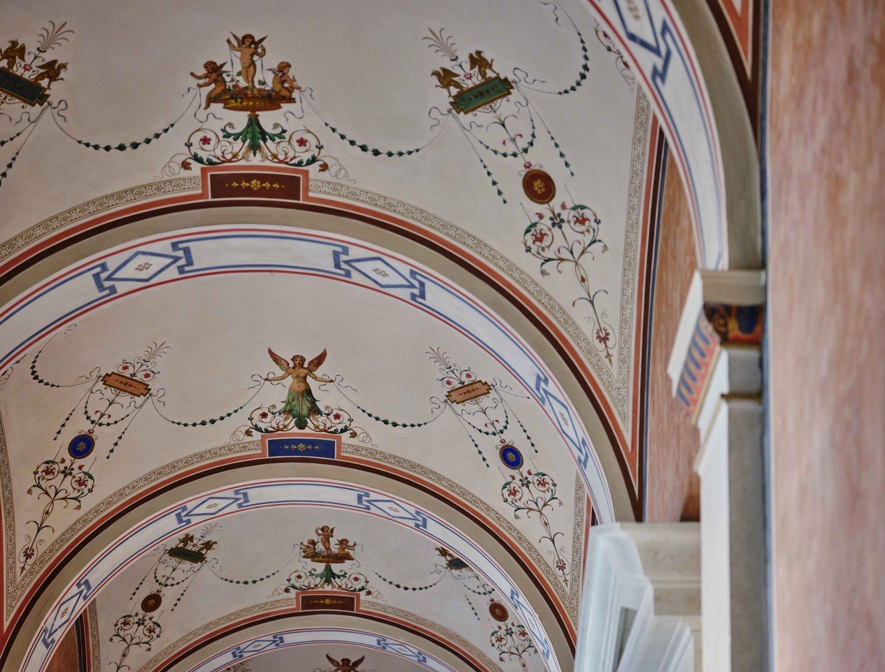 Detail of the ceiling painting in the vault of the academy building, this is framed by geometric shapes, in the vault small golden putti standing together in groups of three on a pedestal holding green plant garlands, under the scene a fine ornamentation of red-green ornaments