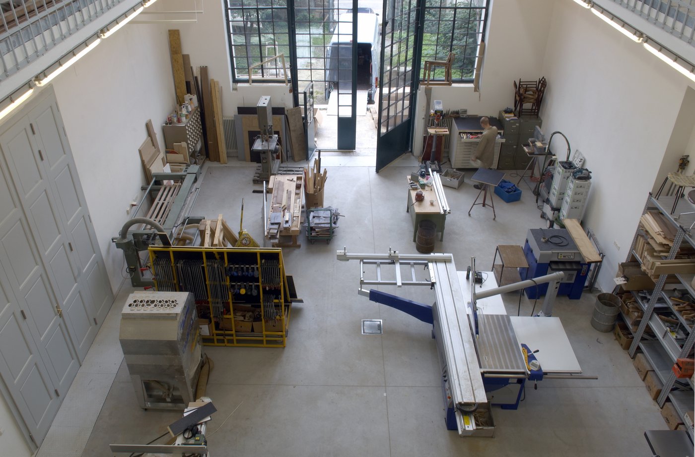 View into the workshop from above, you can see many tools for woodworking, shelves and wooden planks. In the background, the gate opens to the garden, where there is a van with an open loading area.