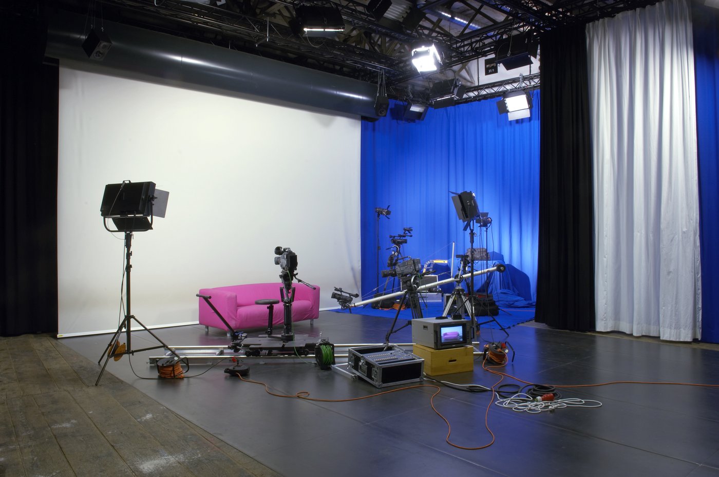 A typical video studio with a variety of cameras, monitors and other equipment. A pink sofa, on the back wall a blue curtain.