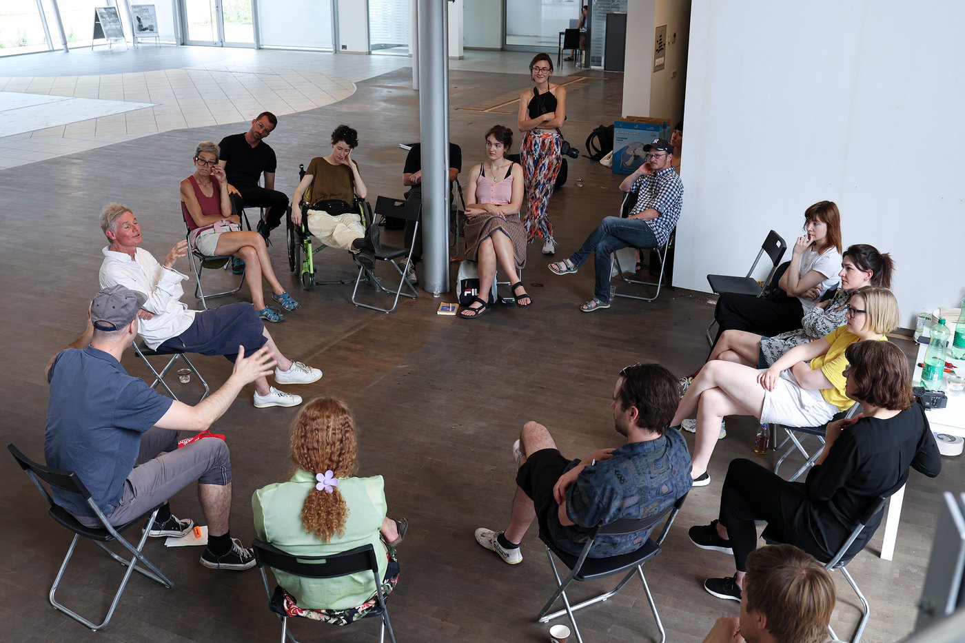 A large group of people sit on armchairs in a circle and listen, a man speaks
