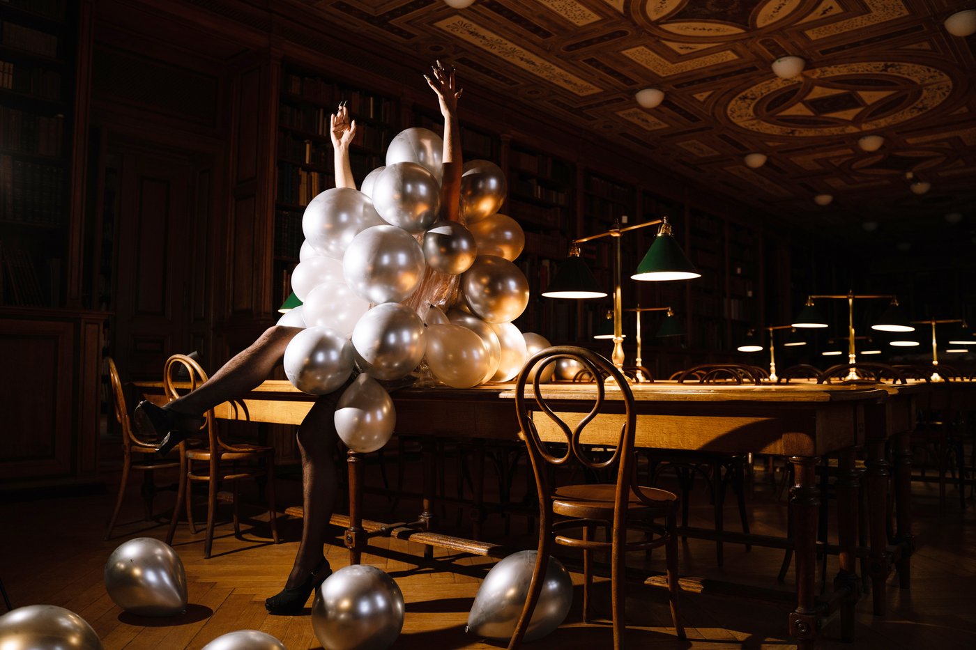 A person wrapped from head to knees in silver balloons sits on a table in the reading room. Except for the glow of the green lampshades, the room is dark. The person is wearing black high heels, one leg is raised. The arms with pointed long fingernails point upwards.