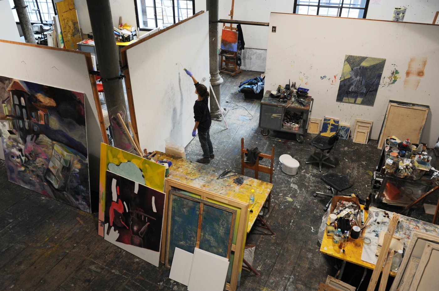 Workplace of a painter photographed from a gallery. Stacks of half-finished canvases stand around, tables with paints and brushes. The floor is littered with splashes of paint. A student in black clothes stands in front of a white wall and draws.