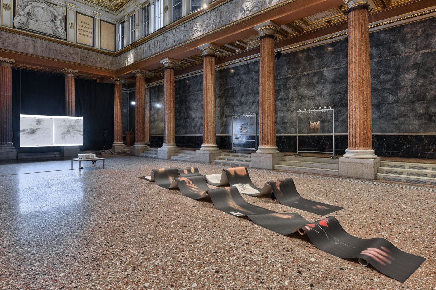 Printed paper webs are laid out on the marbled floor of the auditorium. You can see various projections and an installation with coat racks. The portico of the auditorium and the frieze are also visible.