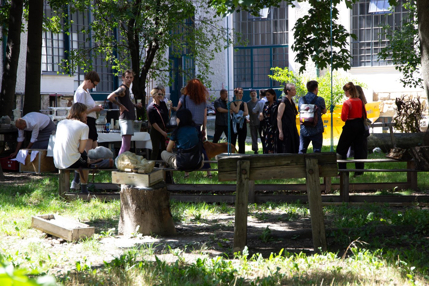 People stand at a solemn gathering in a green courtyard. It is summer, benches and a table with drinks have been set up. Between them are equipment and boxes, parts of sculptures. In the background you can see the large studio windows of the building.