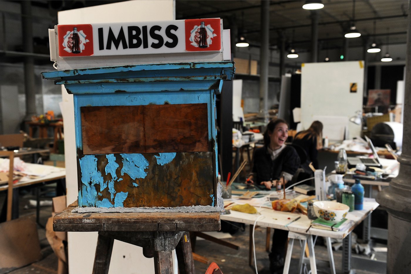 In the foreground you can see the model of a stage setup, a kiosk with peeling blue paint and the inscription "Imbiss" above it. In the background you can see the studio situation with students at tables filled with working material.
